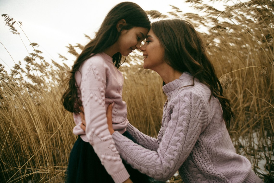A mother and daughter wear matching sweaters and look at each other lovingly in a field of wheat