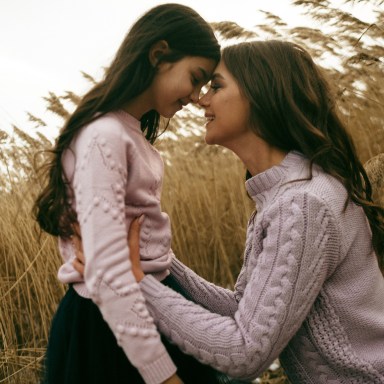 A mother and daughter wear matching sweaters and look at each other lovingly in a field of wheat