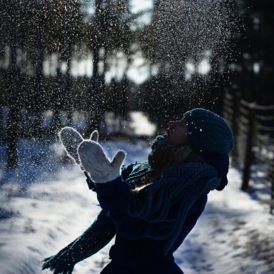 A girl bundled up in winter clothes happily plays with snow while standing on a snowy path