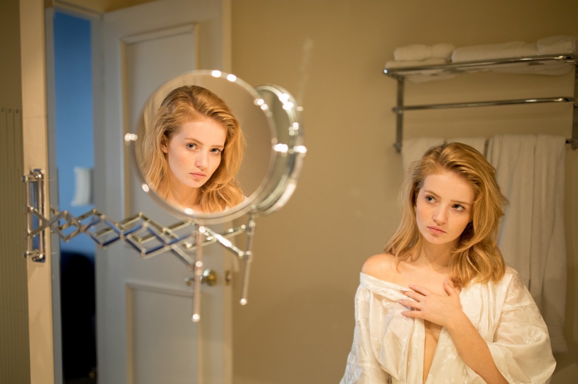 A woman in a white robe looks at herself in a mirror; her reflection stares at the camera