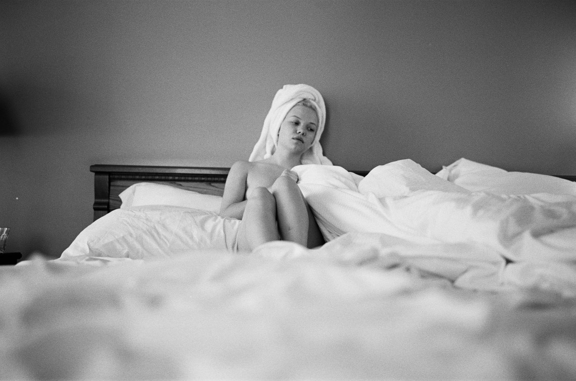 A woman sits in bed half-naked while being surrounded by blankets
