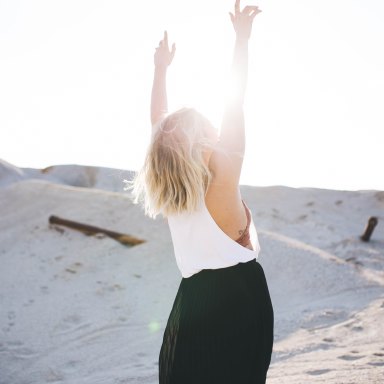 Woman walks in the sand and throws her hands up into the sun