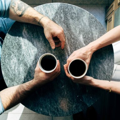 A man and a woman sit across from each other at a table, holding coffee