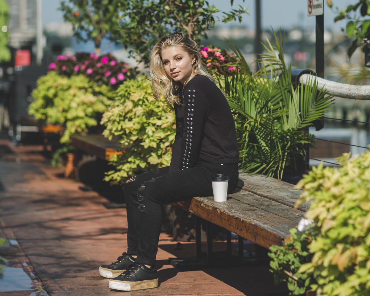 blond woman dressed sitting on a park bench with a paper cup beside her