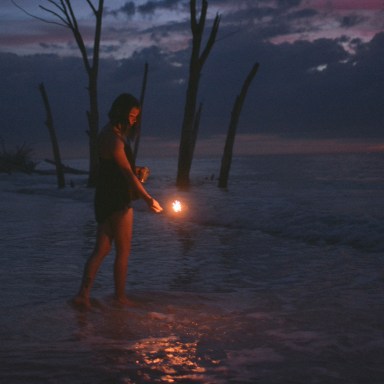 Girl playing with fire, sparkler standing in water with purples and blues of sunset reflecting, dead trees