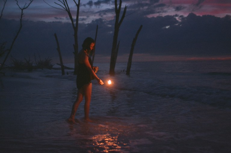 Girl playing with fire, sparkler standing in water with purples and blues of sunset reflecting, dead trees