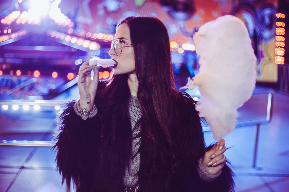 Goth girl eating fluffy pink cotton candy in her black furry jacket at an amusement park