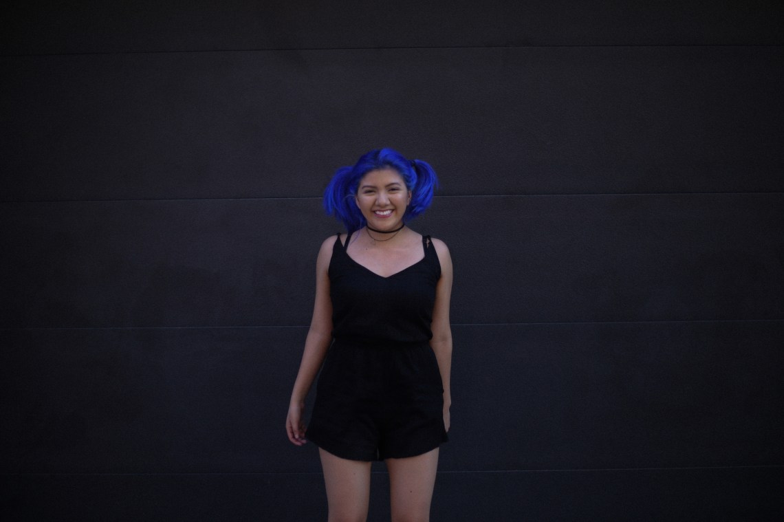 A woman with blue hair stands staring at the camera while she stands in front of a wall