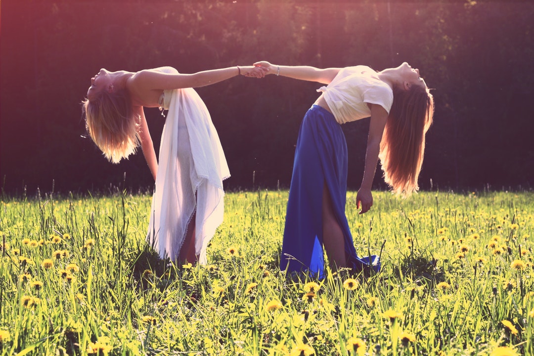 Two women in dresses arching back while holding each other hands in a green meadow