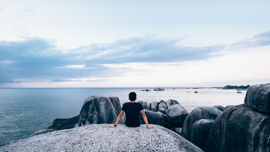 A man sits on a rock, overlooking rocks and the ocean, into the horizon.