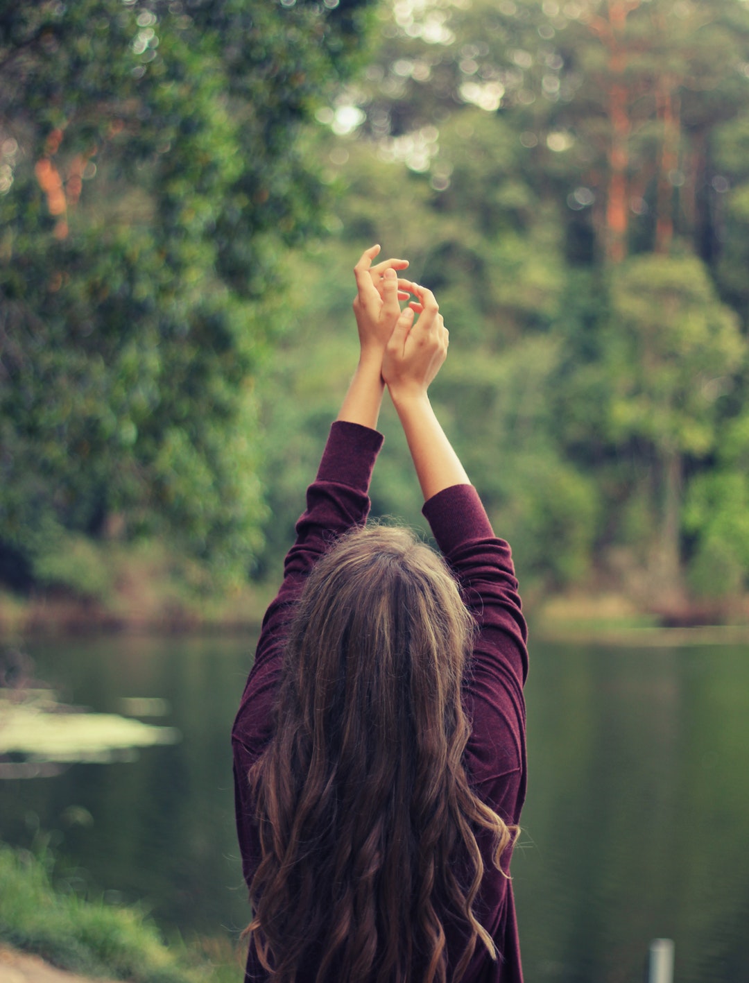 A woman in a purple sweater holds her arms in the air outdoors aside a lake