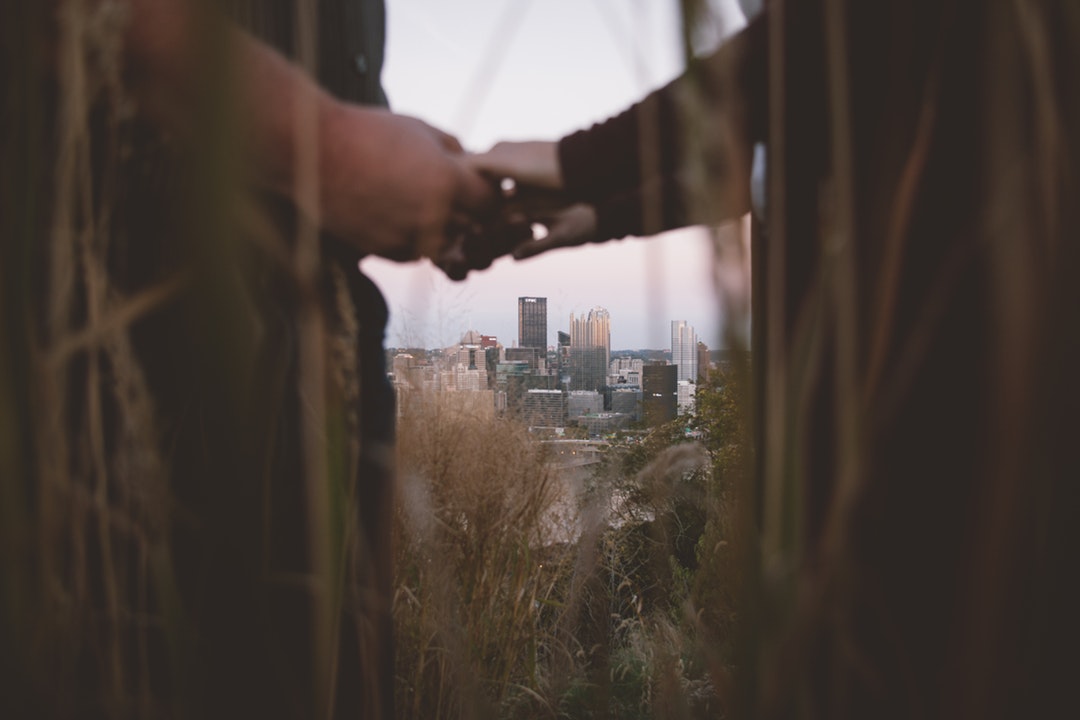 A couple is seen holding hands through tall grasses, with the Pittsburgh skyline in the background