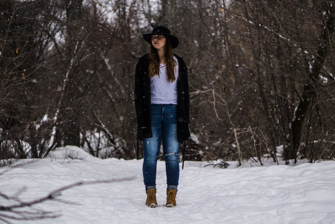A woman in a hat stands in a wintry forest as snow falls around her