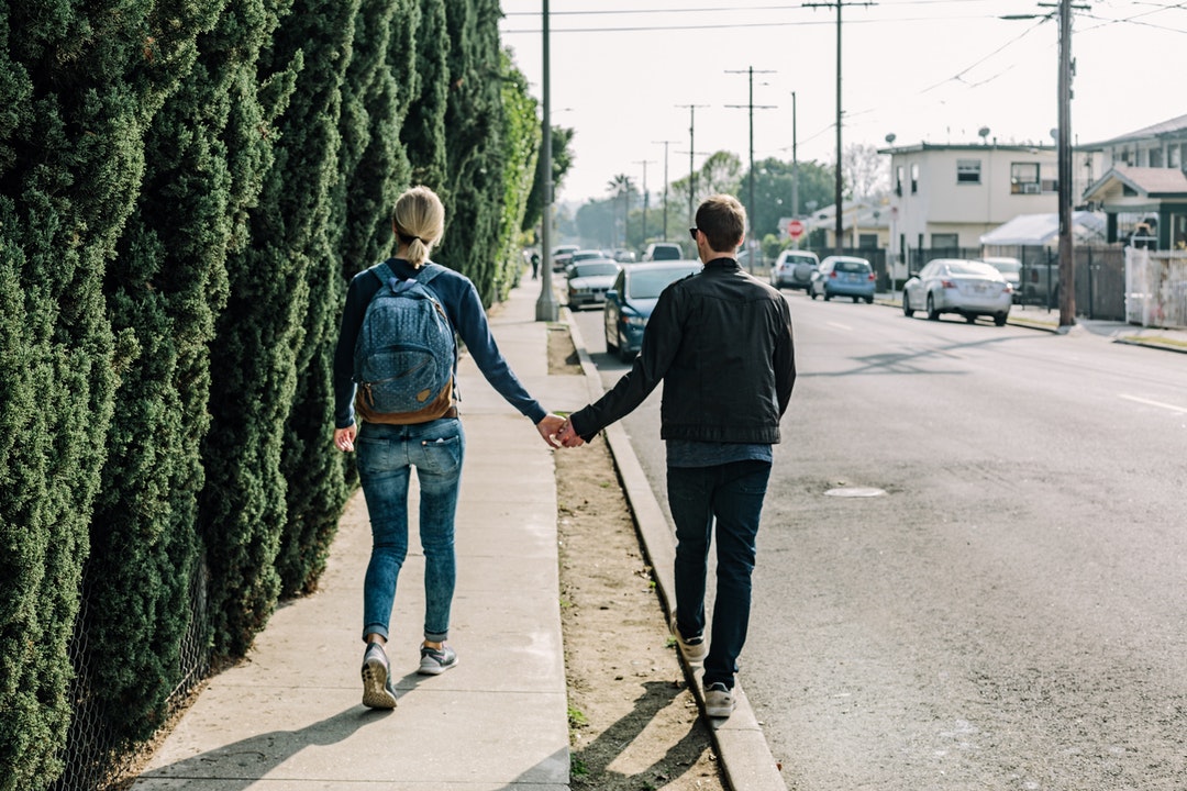 A couple walks holding hands, one on the sidewalk and one on the curb
