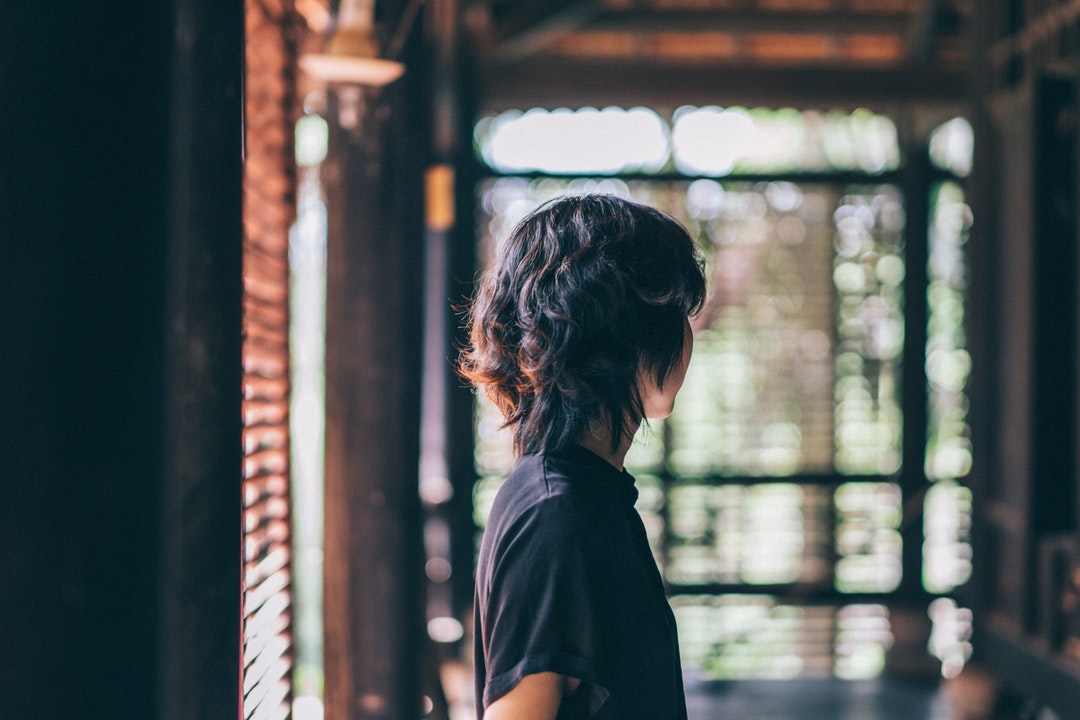 A person stands in a hallway facing away toward a window with open blinds