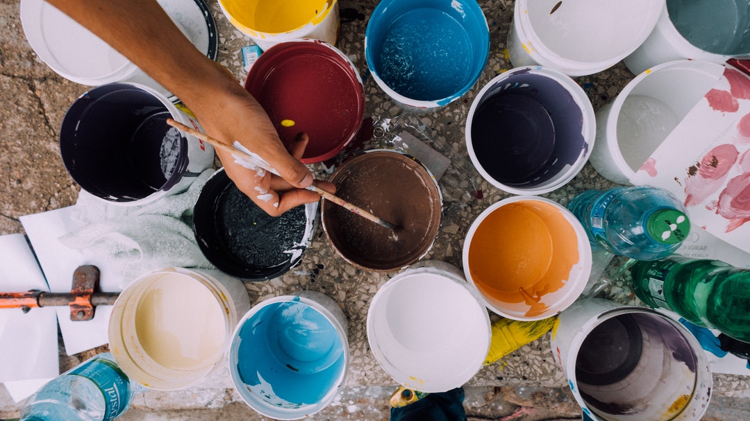 Artist dips paintbrush in bucket of paint among other pots of different colored paint