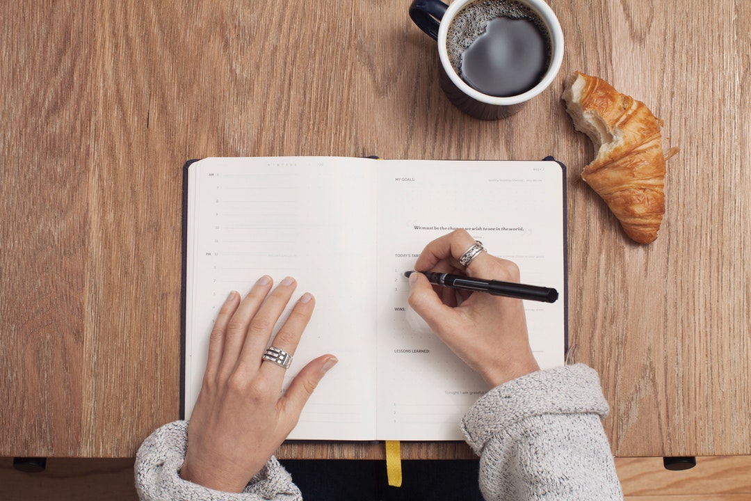 A person writing in a life planner with a coffee and a croissant on the desk