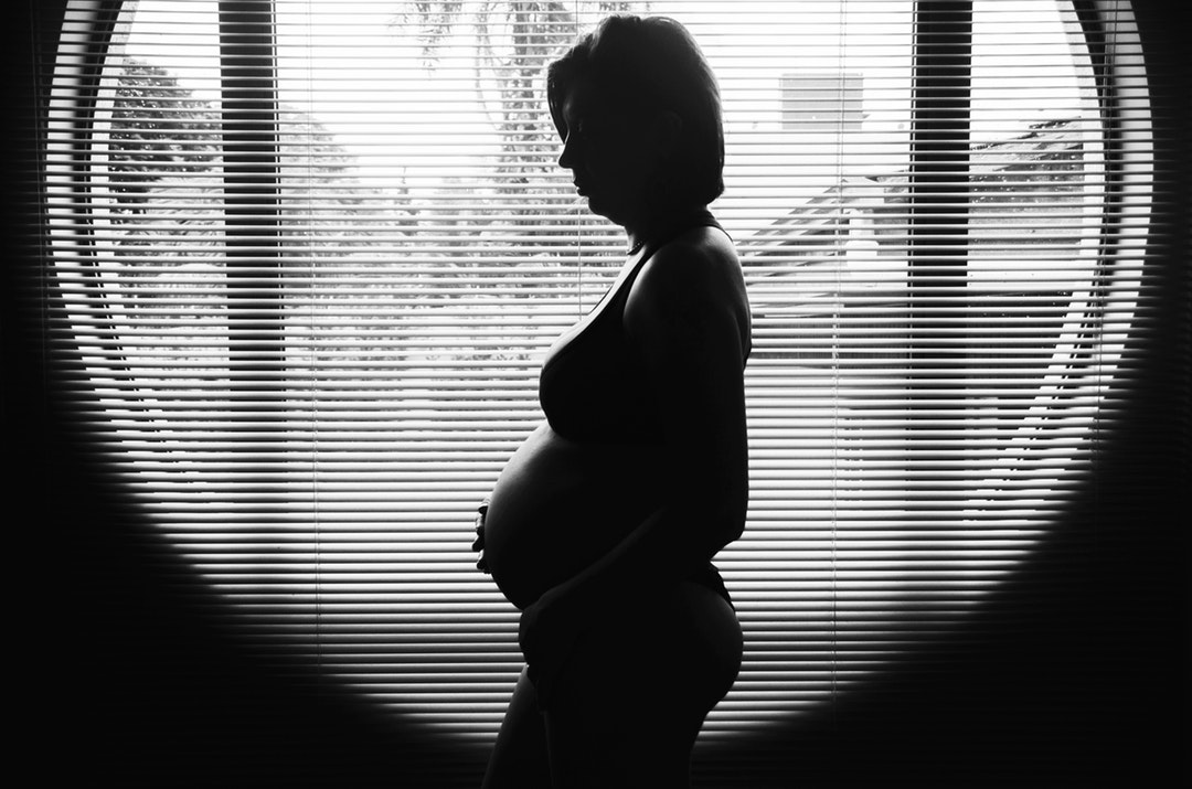 The black and white silhouette of a pregnant woman, standing in front of a round window.