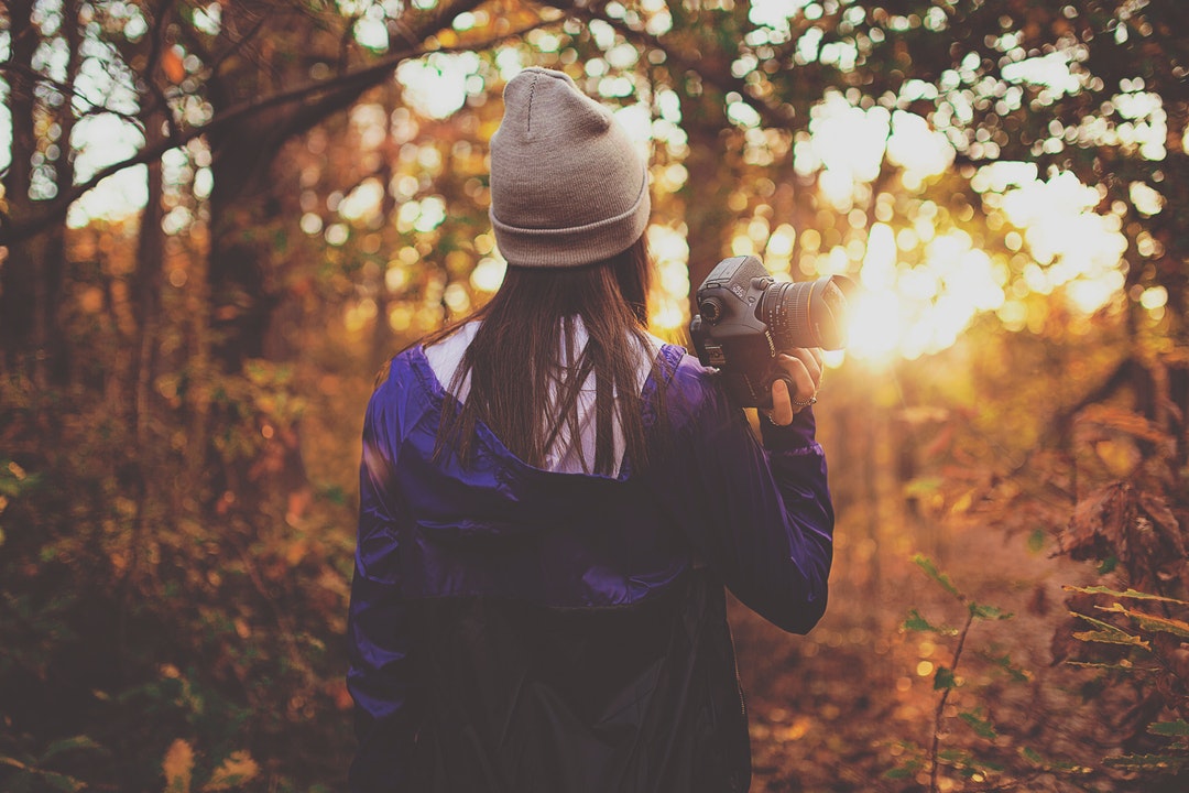 A woman in a beanie walking through a forest with a camera in her hand during sunset