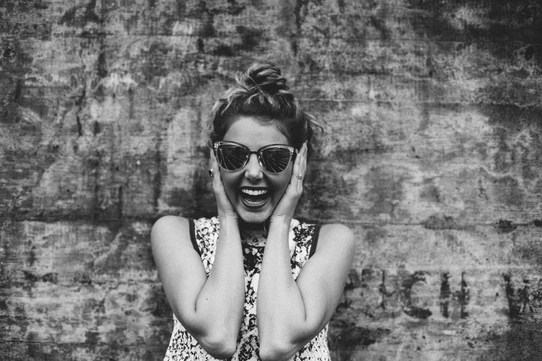A woman with a large smile wearing sunglasses with her hands up on either side of her head