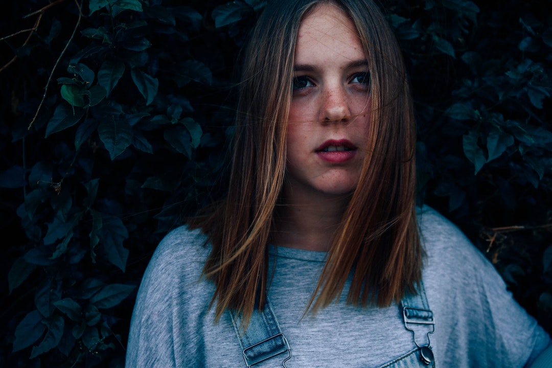 A woman with a gray shirt and denim overalls stands in front of a leafy wall
