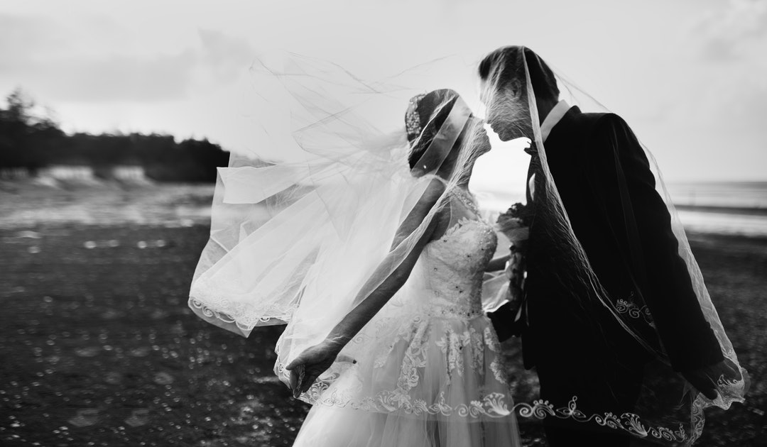 Black and white photo of a bride's veil flying up over her new husband as they kiss