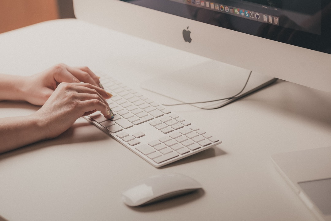 A woman's hands on a computer keyboard on a white workspace with an iMac computer