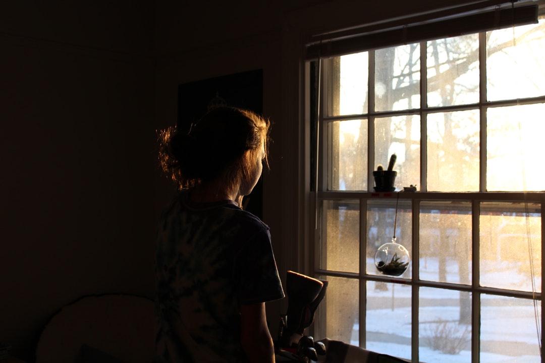 A woman's dark silhouette looks out the window into the brightness of the day.