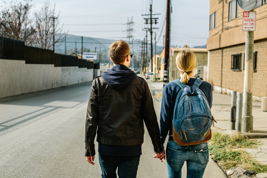 A man and woman wearing a backpack walk along a city street holding hands