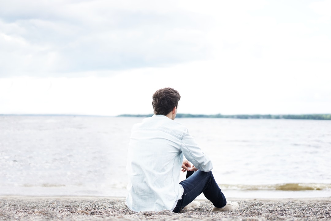 A man in a white shirt sitting on a pebble beach and looking of into the sea
