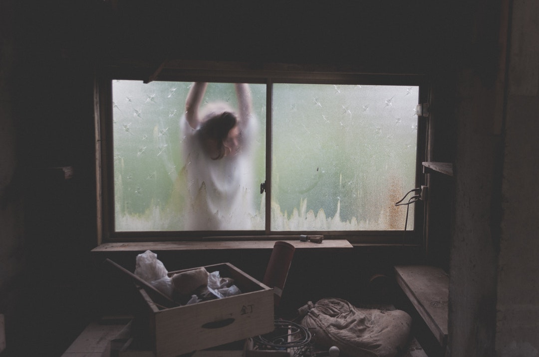 A spooky shot of a helpless looking woman clawing at a window from the outside, with the interior of the room covered in boxes