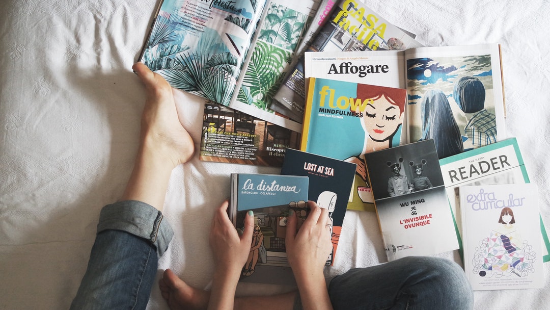 A woman sitting on her bed in jeans looking at several books and magazines in Milan