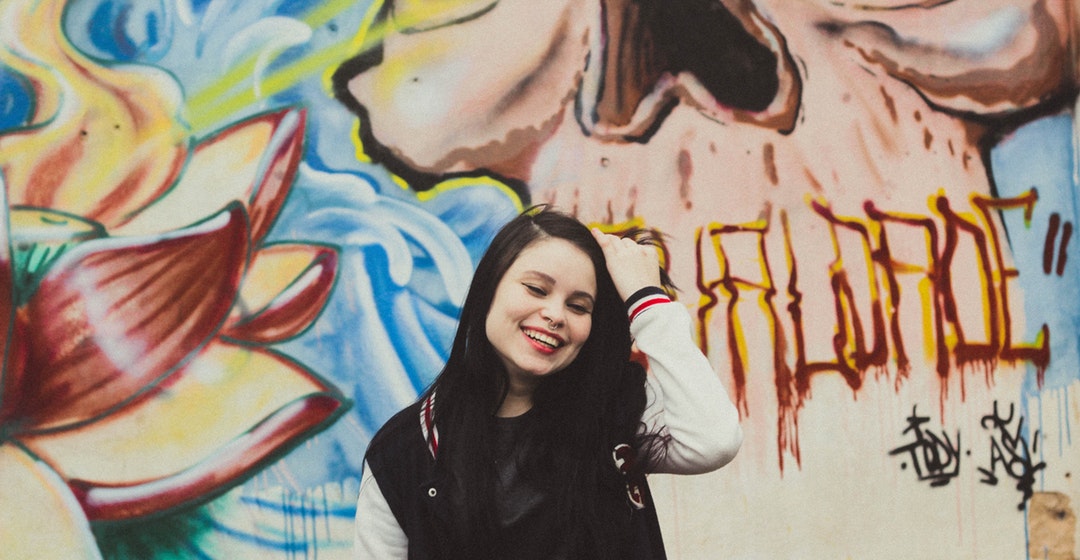 Young smiling hipster girl in baseball jacket in front of colorful graffiti mural