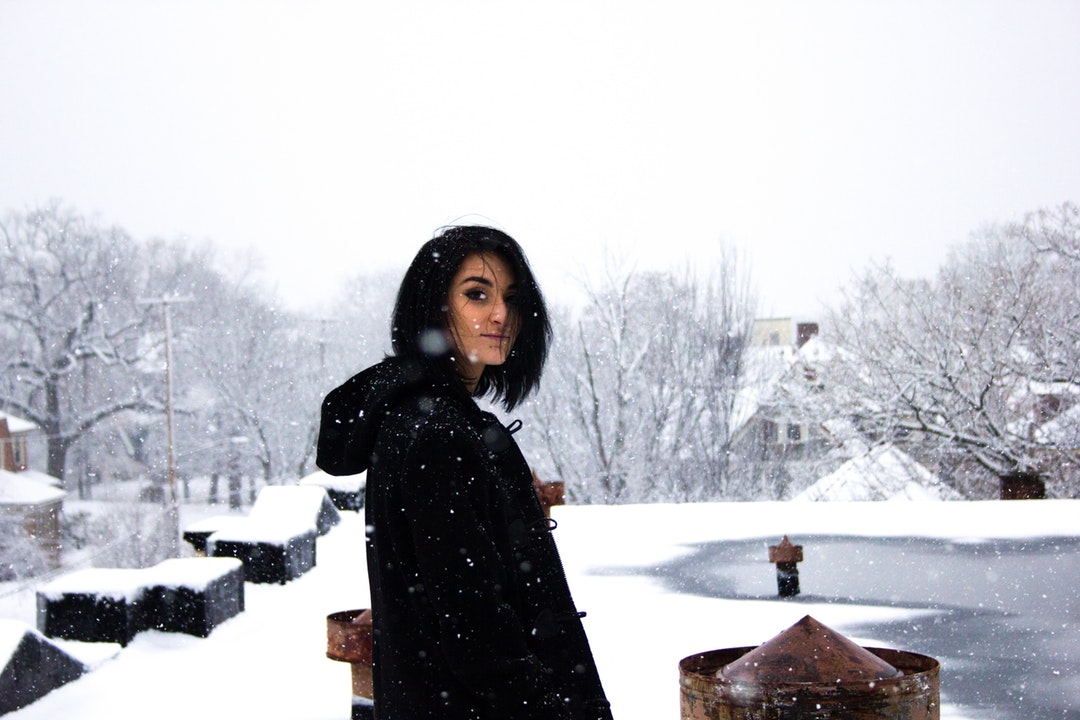 A dark-haired woman in a black coat on a snowy roof of looking back