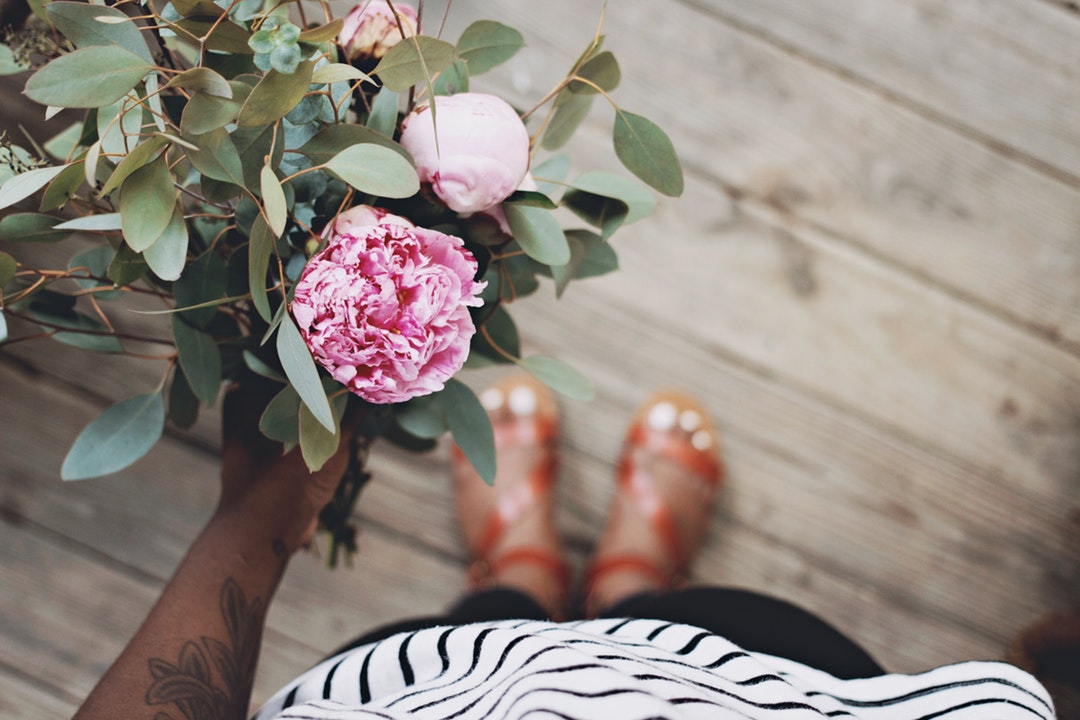 Woman with flower tattoos and sandals holding a bouquet of flowers while standing on wooden floor