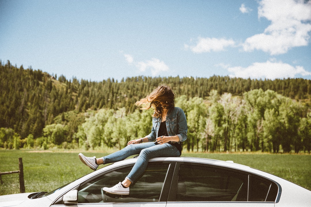 Young woman in denim sitting on white car roof with evergreen forest in background