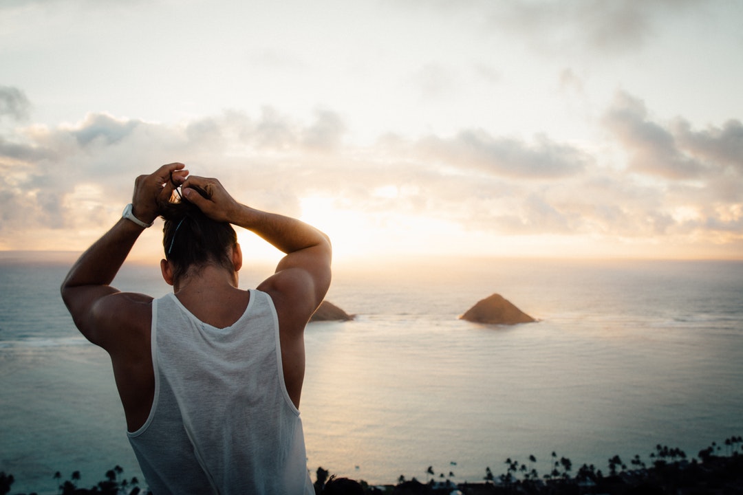 A man standing on a mountain by the coast putting his hair into a bun
