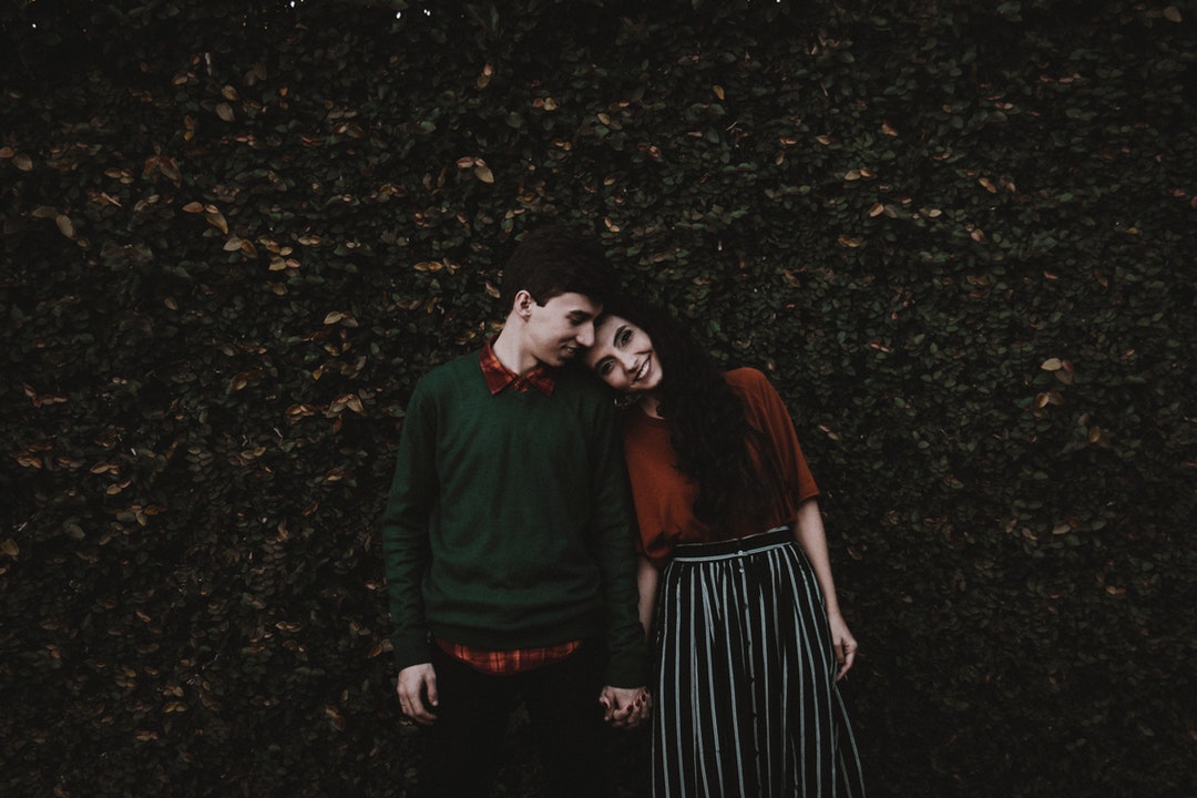 Couple holds hands and leans their heads together in front of a leafy wall in Cianorte