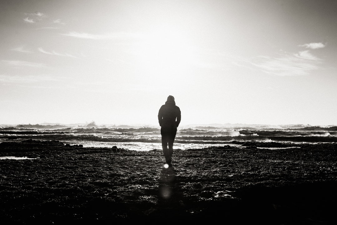 Black and white shot of person walking towards sea water on shore with clouds in sky