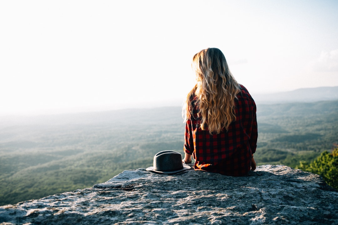 A blonde woman with a hat on her side sitting on a rocky ledge overlooking green plains