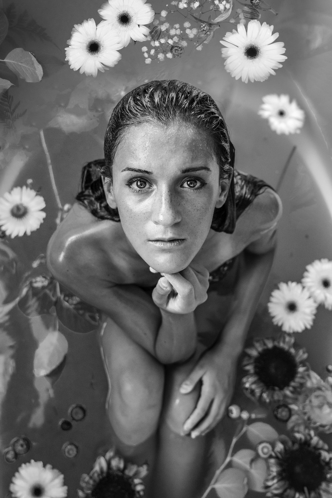 A black-and-white shot of a woman sitting in a bath filled with water and flowers