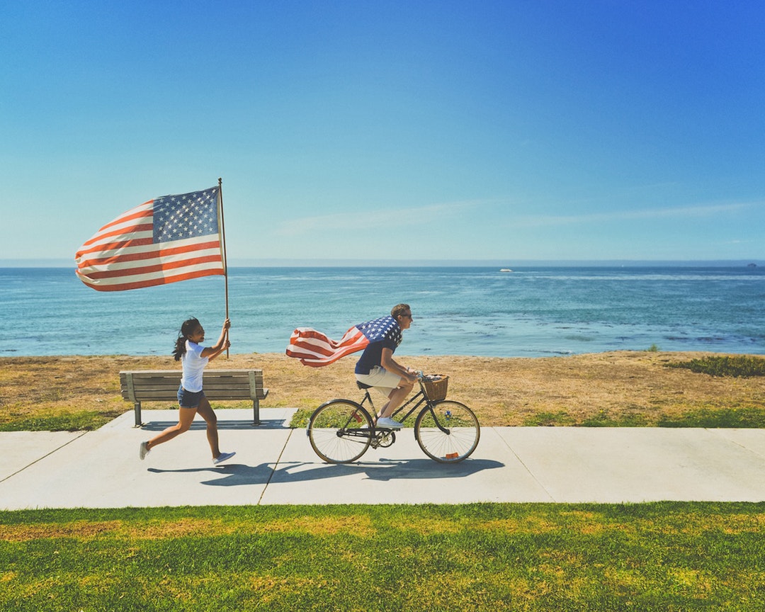 Man on a bicycle followed by a woman running with US flags at Shell Beach shoreline path