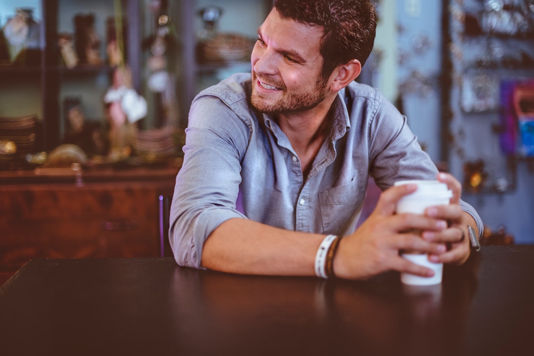 A man in a denim shirt sitting at a table and clutching a cup of coffee