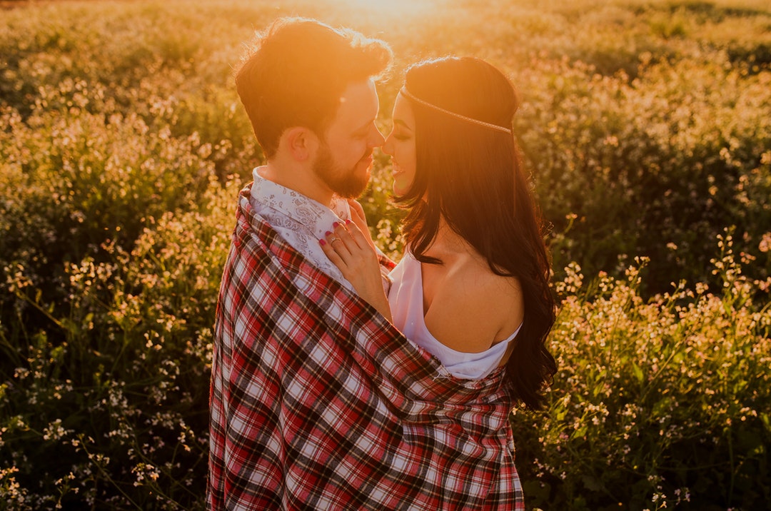 Bearded man and woman in a headband hug, wrapped in a blanket in a meadow