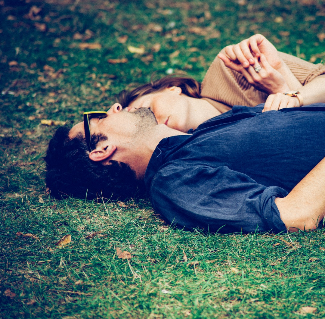 A man in sunglasses and a woman lay together in green grass with scattered fallen leaves