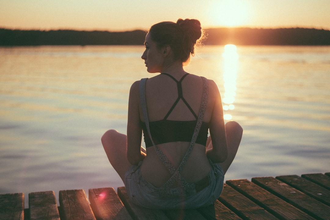 Woman in dungarees sitting on dock looking to the left whil sun sets in the horizon and reflects over the water in Olsztyn