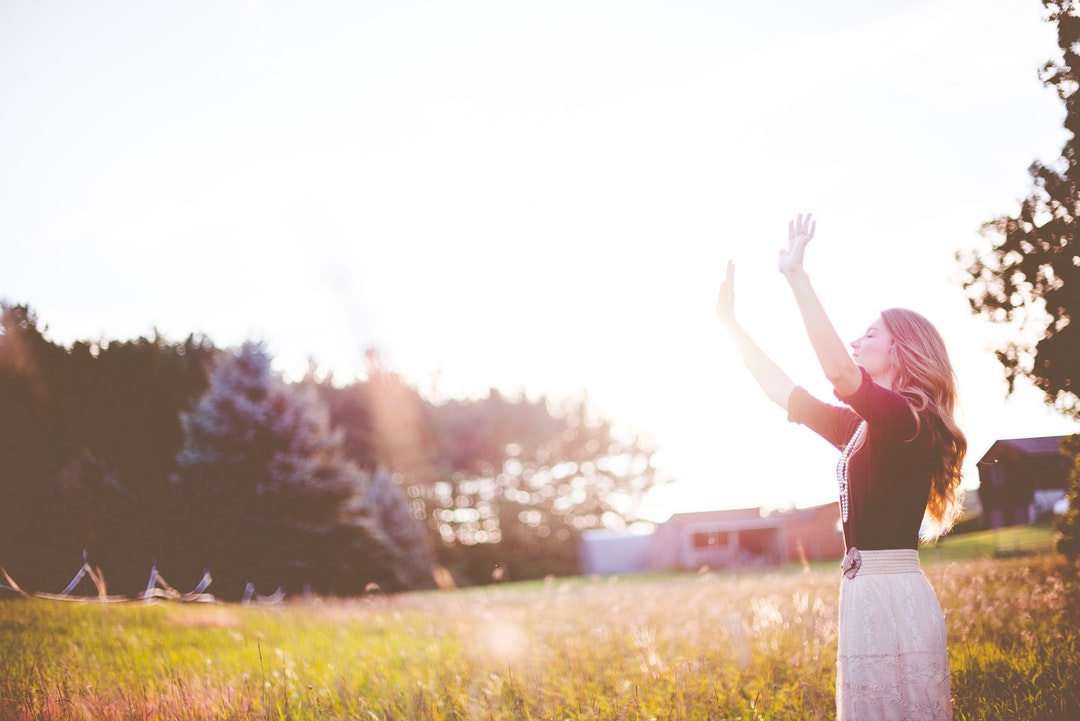 A blonde woman in a field raising her hands to the sky.