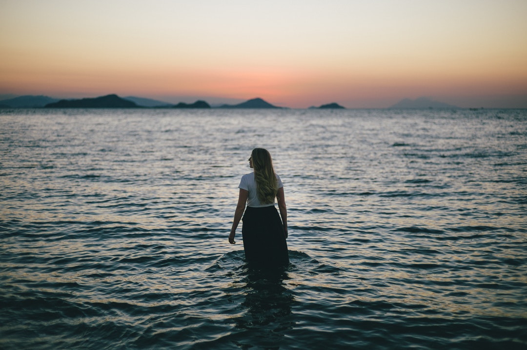 The back of a woman standing knee-deep in the ocean looking into the distance at dusk