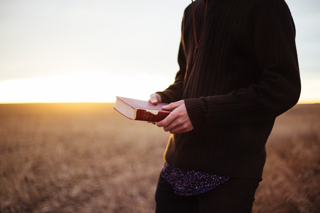 Man in sweater holding bible with two hands in a field during sunset