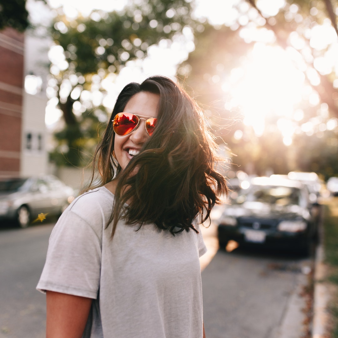 A smiling woman in sunglasses and a t-shirt stands on a sunny street in Chicago